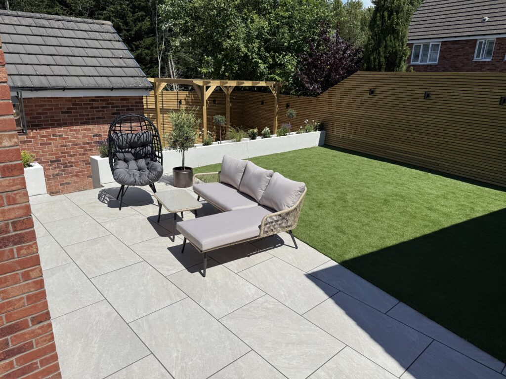 Contemporary patio with light-grey porcelain paving, outdoor lounge seating, raised planters, artificial lawn, and a timber pergola.