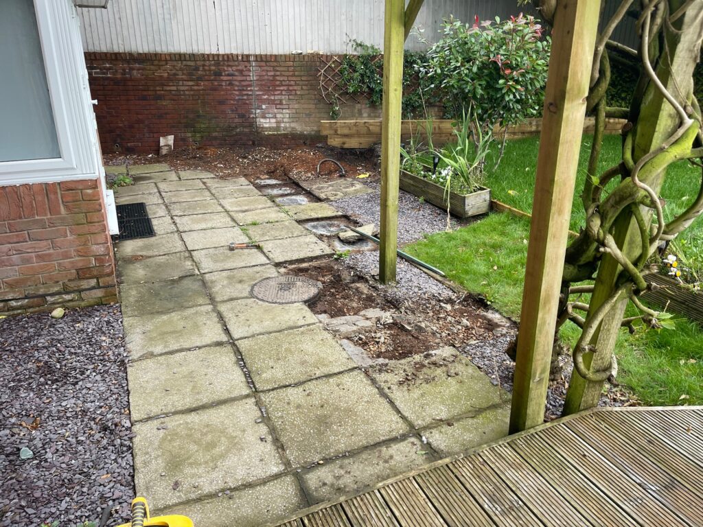 Before garden transformation showing worn concrete paving, uneven ground and outdated patio area beside residential property