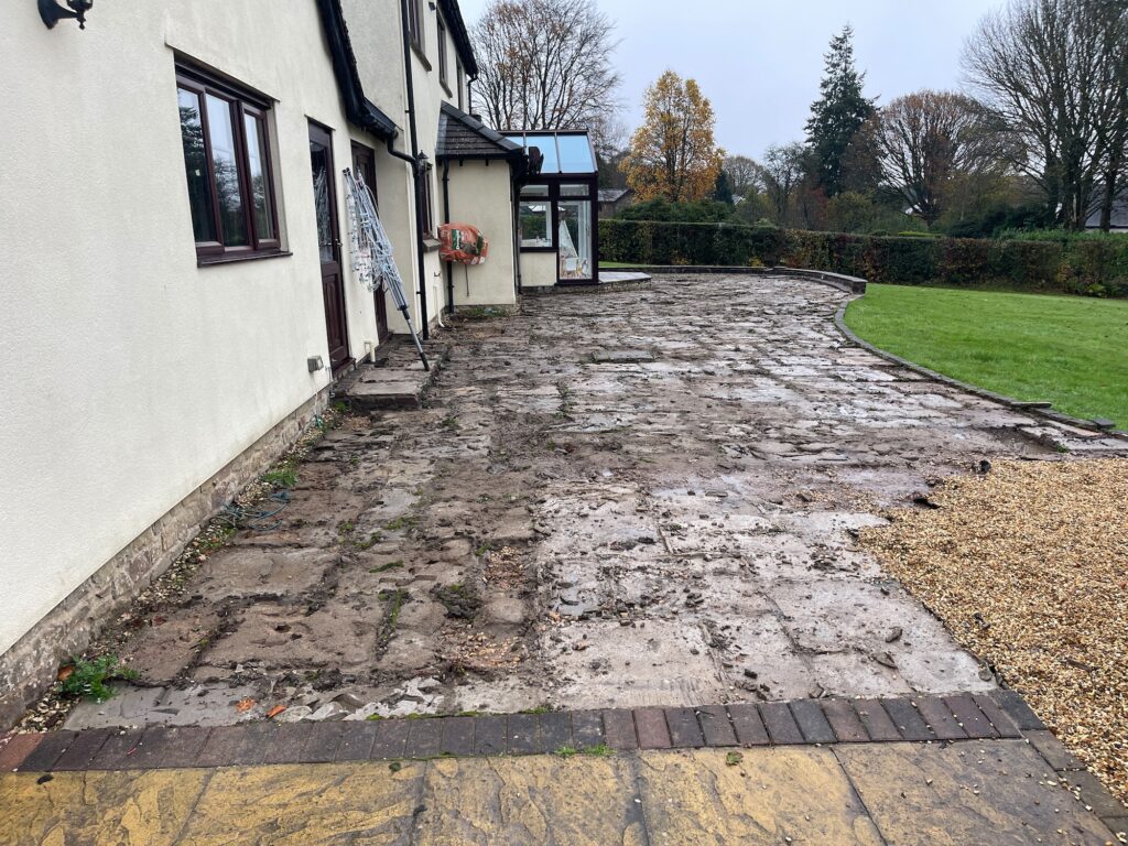 Before garden transformation showing worn and uneven patio paving with muddy surface and poor drainage around large residential property